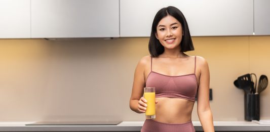 Jeune femme japonaise mince et souriante qui boit un jus de fruits dans une cuisine en train de cuisiner un dîner sain