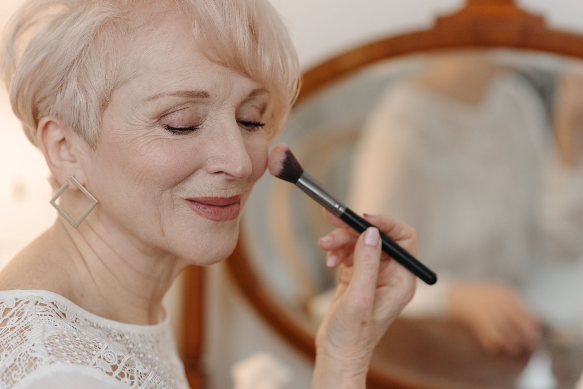 Femme âgée aux cheveux courts blonds appliquant du blush sur la joue avec un pinceau, devant un miroir ovale à cadre en bois.