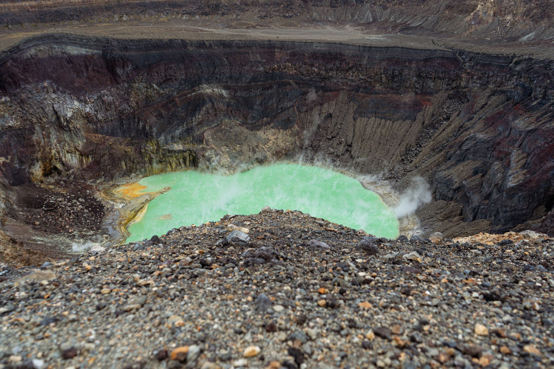 Lac vert turquoise fumant au fond d’un cratère volcanique, entouré de parois rocheuses et de gravier au premier plan.