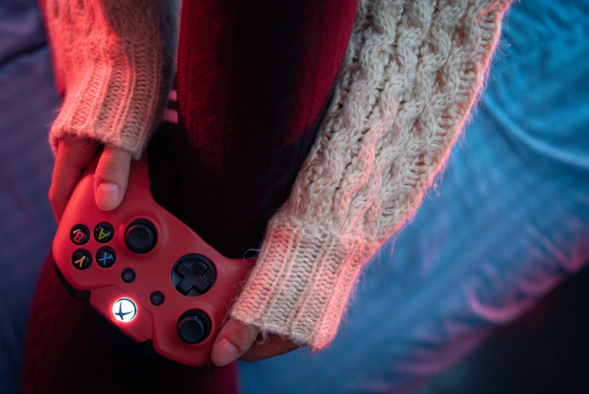 Mains aux manches de pull en maille beige tenant une manette de jeu rouge au bouton central lumineux, sous un éclairage rose et bleu.