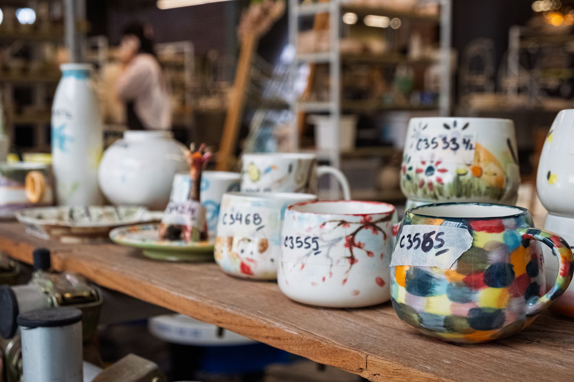 Tasses en céramique peintes à la main, colorées, alignées sur une table en bois dans une boutique, avec des étiquettes à codes manuscrits.