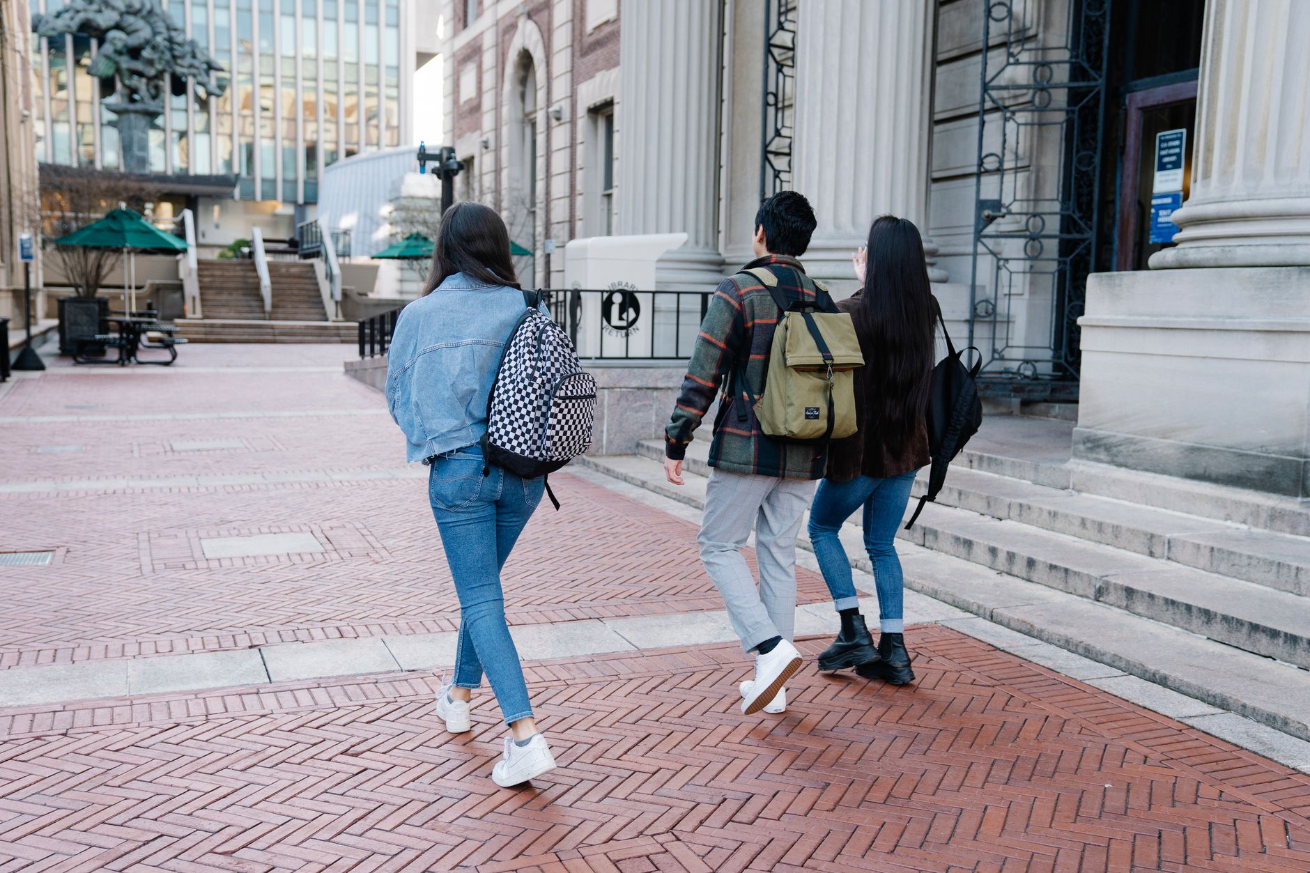 Trois étudiants portant des sacs à dos marchent de dos sur une place pavée près d’un bâtiment à colonnes.