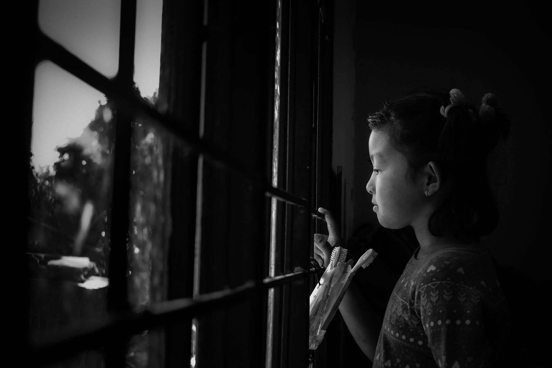 Une enfant de profil regarde par une fenêtre à petits carreaux dans une pièce sombre, une main contre la vitre, en noir et blanc.