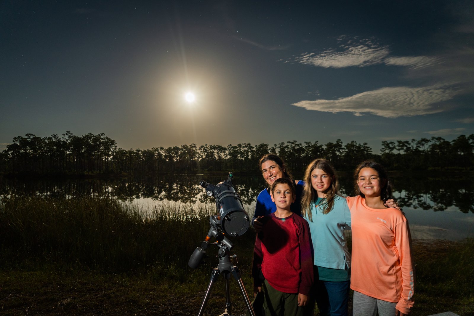 Une femme et trois enfants posent près d’un télescope sur trépied, de nuit, au bord d’un plan d’eau, sous un ciel éclairé par la lune.